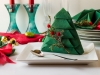 Green color,paper napkin folded in Christmas tree shape on white surface with dinnerware and ornaments.